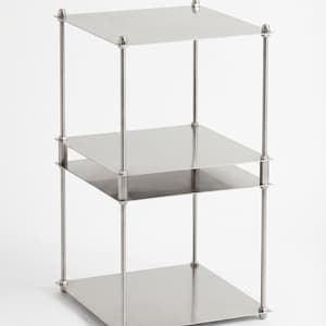 May include: A three-tiered, stainless steel shelf unit. Each square shelf is supported by four vertical posts with decorative finials at the top and bottom. The unit is designed with a minimalist aesthetic and a polished finish.