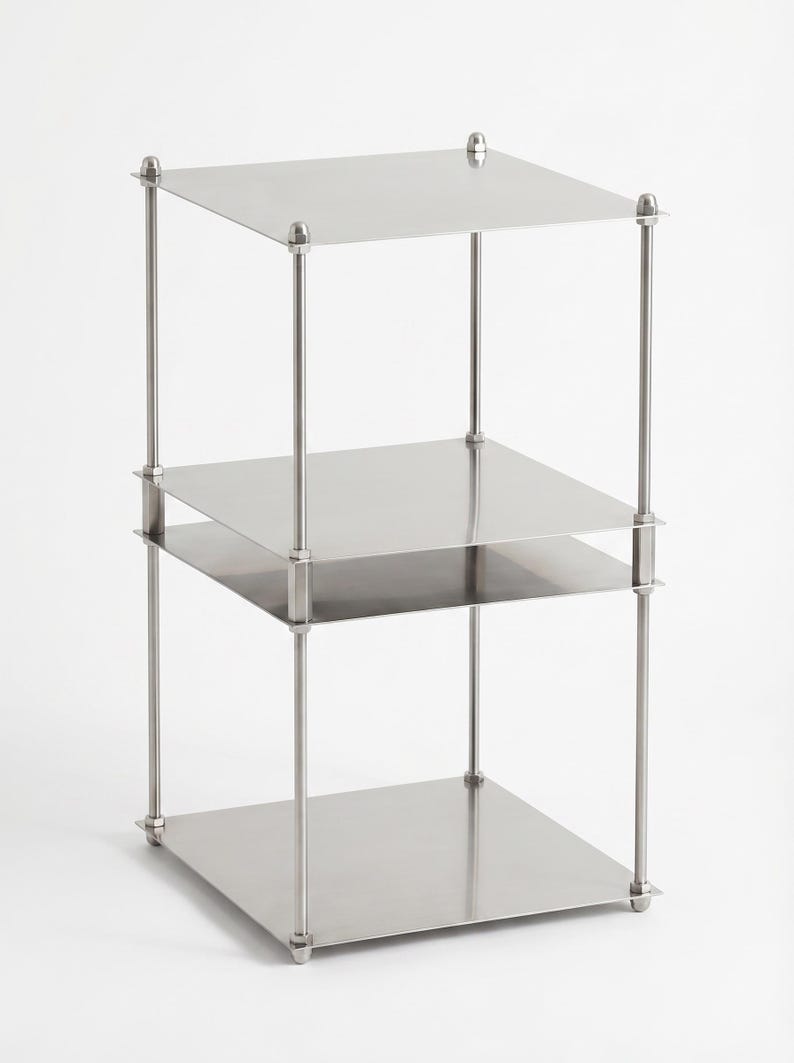 May include: A three-tiered, stainless steel shelf unit. Each square shelf is supported by four vertical posts with decorative finials at the top and bottom. The unit is designed with a minimalist aesthetic and a polished finish.