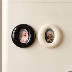 May include: Two oval-shaped magnetic picture frames, one in glossy black and one in cream, are attached to a cream-colored surface. Each frame holds a small photograph of a light-colored, fluffy dog with a small red accessory on its head.