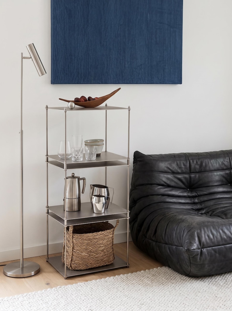 May include: A modern living room scene featuring a stainless steel shelving unit with various items. The shelves hold glassware, a coffee pot, a pitcher, and a woven basket. A black leather sofa and a floor lamp are also visible.