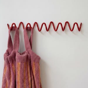 Pode incluir: Um cabide de parede vermelho brilhante com um design de fio metálico contínuo em forma de ziguezague ou onda. Duas toalhas de felpo às riscas cor-de-rosa e laranja com alças de tecido estão penduradas nos ganchos do suporte metálico.