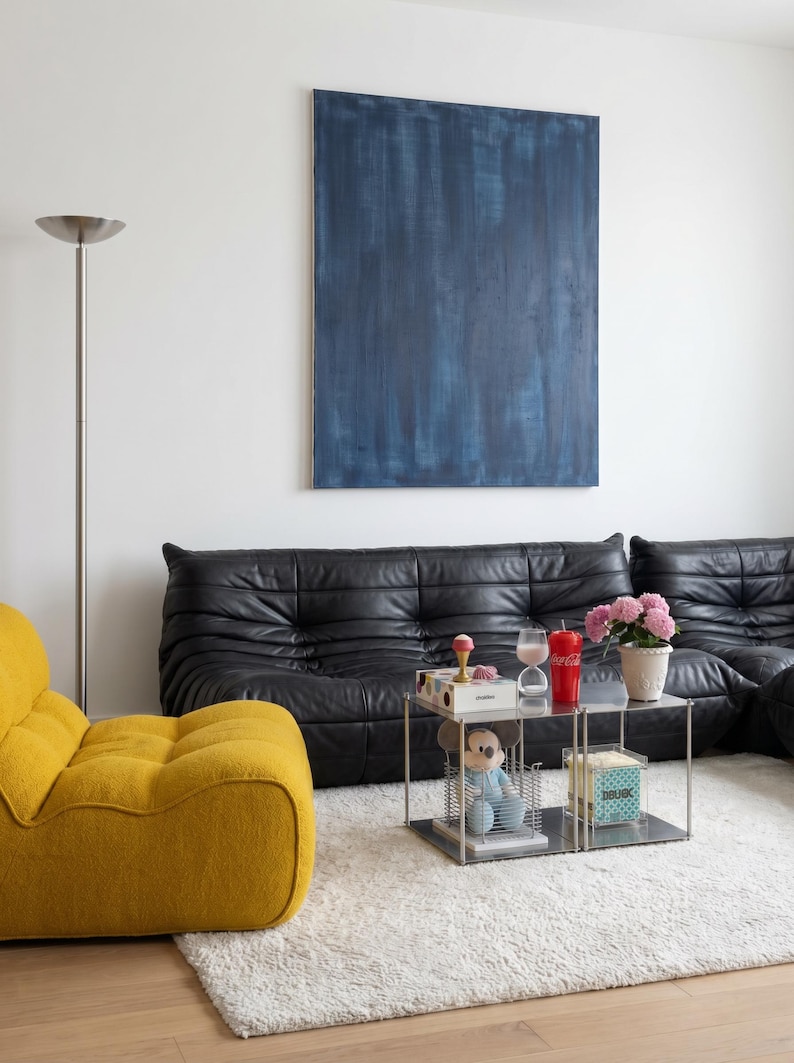 May include: A modern living room with a black leather sofa, a yellow armchair, and a small, square, metal coffee table. A large blue abstract painting hangs on the white wall. The room has a white rug and a floor lamp.