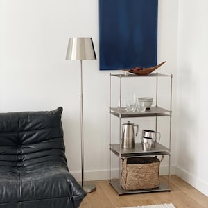 May include: A modern living room scene featuring a silver floor lamp, a black leather armchair, and a metal shelving unit. The shelves hold glassware, a coffee pot, and a woven basket. A blue painting hangs on the wall.