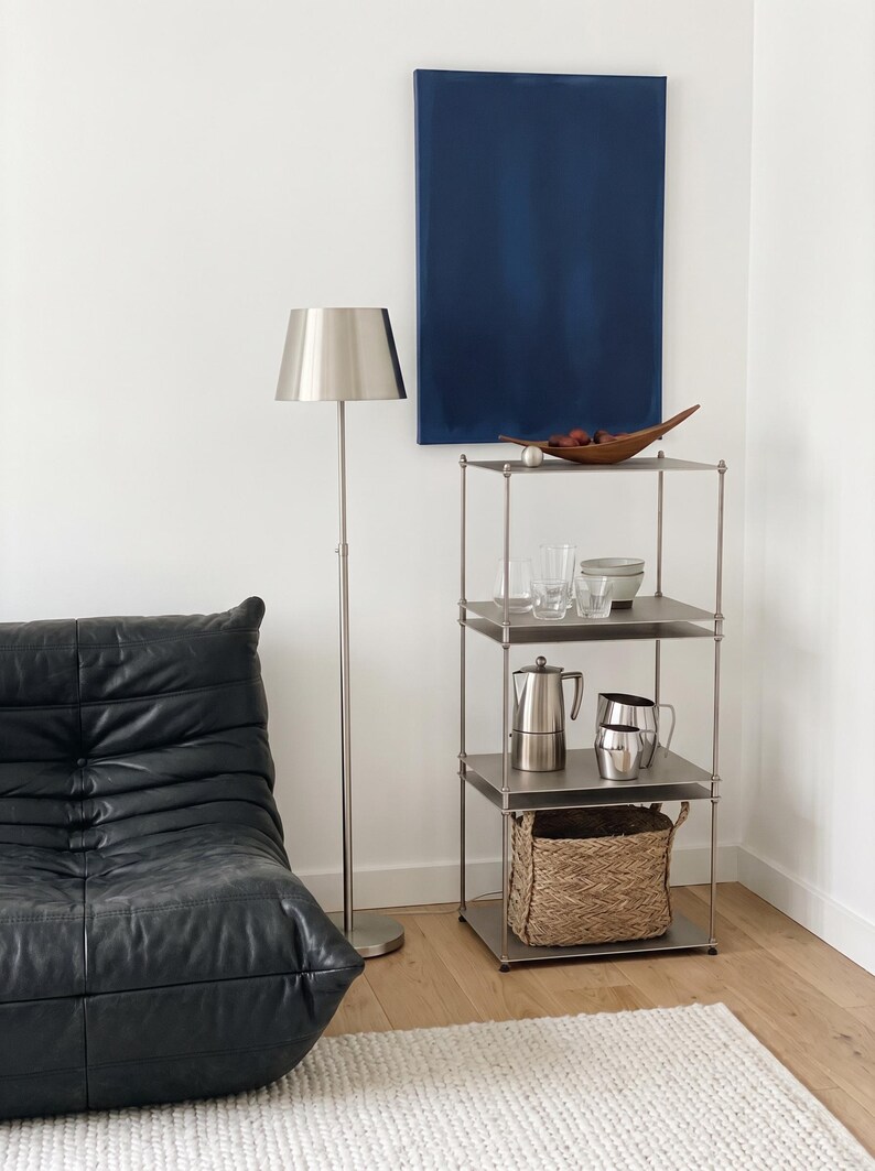 May include: A modern living room scene featuring a silver floor lamp, a black leather armchair, and a metal shelving unit. The shelves hold glassware, a coffee pot, and a woven basket. A blue painting hangs on the wall.
