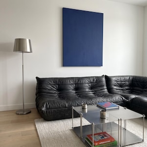 May include: A modern living room featuring a black leather sectional sofa, a stainless steel coffee table with books, and a silver floor lamp. A large, solid blue painting hangs on the white wall. The room has light wood flooring and a white rug.