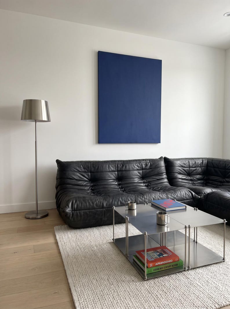 May include: A modern living room featuring a black leather sectional sofa, a stainless steel coffee table with books, and a silver floor lamp. A large, solid blue painting hangs on the white wall. The room has light wood flooring and a white rug.
