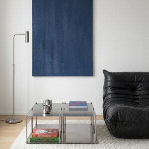 May include: A modern living room featuring a large blue abstract painting, a stainless steel coffee table with books and mugs, a black leather sofa, and a silver floor lamp. The room has a minimalist design with a white rug.