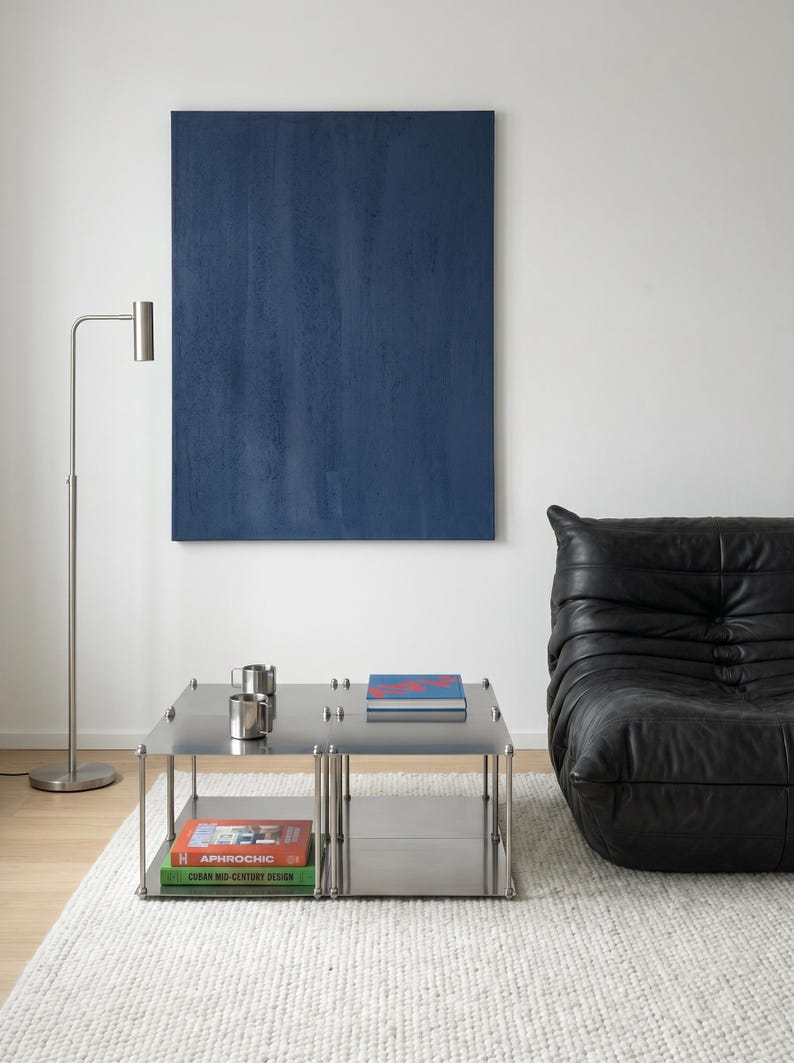 May include: A modern living room featuring a large blue abstract painting, a stainless steel coffee table with books and mugs, a black leather sofa, and a silver floor lamp. The room has a minimalist design with a white rug.