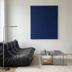 May include: Modern living room with a black leather lounge chair, a stainless steel coffee table, and a large blue painting on a white wall. A silver floor lamp and a white rug complete the contemporary design.