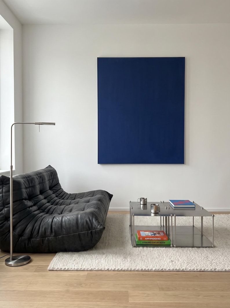 May include: Modern living room with a black leather lounge chair, a stainless steel coffee table, and a large blue painting on a white wall. A silver floor lamp and a white rug complete the contemporary design.