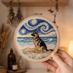 May include: A circular embroidery hoop containing a fabric panel with a hand-stitched scene of a German Shepherd dog sitting on a sandy beach. The background features a swirling, starry night sky in shades of blue and yellow above a turquoise ocean.