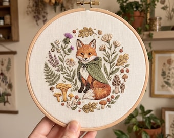 May include: A circular embroidery piece in a wooden hoop. It depicts a fox in a green cape, surrounded by flowers, mushrooms, and leaves. The fox has orange and white fur. The embroidery is on cream fabric, with a wooden hoop.