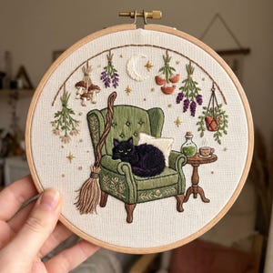 Puede incluir: Obra de arte bordada en un aro de madera. Un gato negro descansa sobre un sillón verde, con una escoba, hierbas y una luna creciente. Una pequeña mesa sostiene una botella de poción y una taza de té. Tema botánico y de brujas.