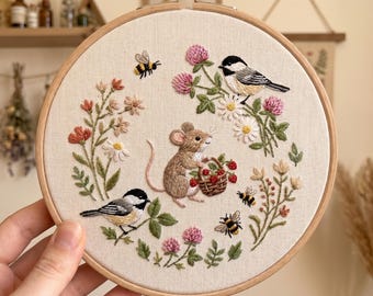 Puede incluir: Obra de arte bordada en un aro de madera. El diseño presenta un ratón que lleva una cesta de fresas, rodeado de pájaros, abejas y flores. La paleta de colores incluye beige, marrón, rosa y verde.