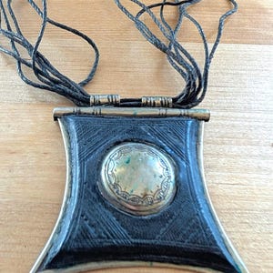 May include: A black pendant necklace with a geometric design. The pendant has a central circular element and is suspended from multiple black cords. The pendant is made of black material with silver-colored metal accents.