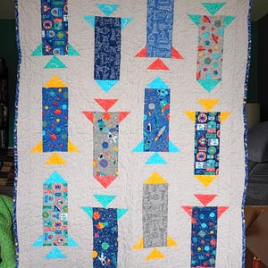 May include: A handmade quilt featuring a repeating pattern of colorful rockets against a light gray background. The rockets have various designs and are accented with teal, red, and yellow triangles. The quilt is bordered with a blue patterned fabric.