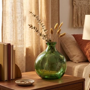 May include: A green glass vase with a round body and a narrow neck, holding dried plants. The vase sits on a wooden surface next to a small, round dish and a stack of books. The background includes a beige curtain and a bed.