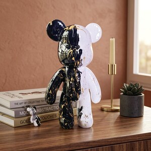 May include: A bear-shaped figurine featuring a split design with one side in glossy black and the other in white, both accented with gold paint splatters. The figure has rounded ears, a smooth surface, and a stylized, anthropomorphic form.