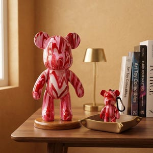 May include: A bear-shaped figurine with a red and white marbled pattern standing on a circular wooden base, accompanied by a smaller matching bear-shaped keychain resting inside a tan leather tray, both featuring a glossy finish and smooth curves.