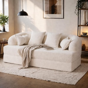 Puede incluir: Un sofá moderno de color crema con brazos redondeados y múltiples almohadas esponjosas. Una manta beige y un libro descansan sobre el sofá. El sofá está sobre una alfombra de color claro en una habitación bien iluminada.