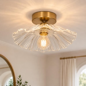 May include: A flush mount ceiling light fixture featuring a circular brushed brass base and a wide, scalloped, clear glass shade with a ribbed texture. A single exposed Edison-style light bulb is centered within the glass, emitting a warm glow.