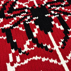 May include: A handmade, textured blanket in a bold pattern of red, black, and white. The design features a central black shape with white and black accents against a red background. The blanket is made of a soft, plush material.