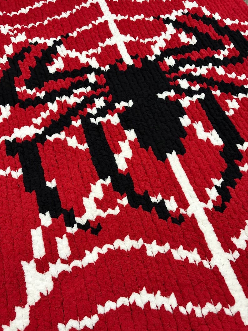 May include: A handmade, textured blanket in a bold pattern of red, black, and white. The design features a central black shape with white and black accents against a red background. The blanket is made of a soft, plush material.