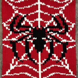 May include: Red knitted blanket featuring a black spider design with a white web pattern. The spider is centered on the blanket, with the web extending outwards. The blanket is made of a thick, textured yarn.