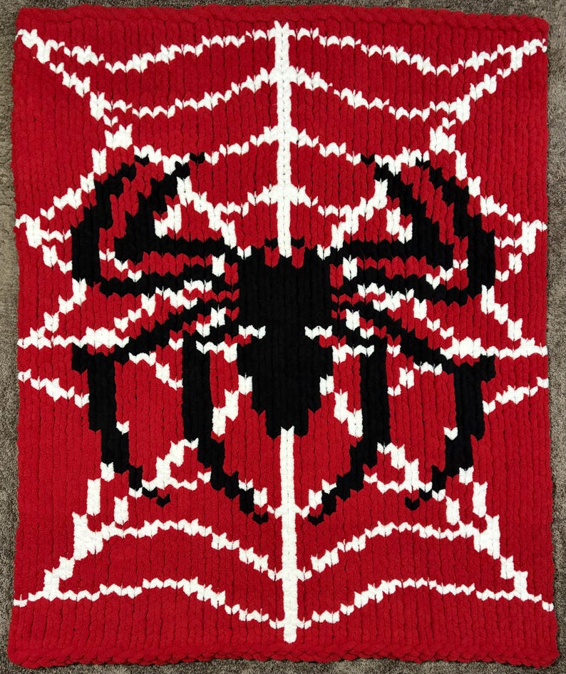 May include: Red knitted blanket featuring a black spider design with a white web pattern. The spider is centered on the blanket, with the web extending outwards. The blanket is made of a thick, textured yarn.