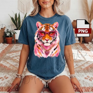 May include: A blue short-sleeve t-shirt featuring a graphic of a tiger wearing pink leopard-print glasses and a gold chain with a heart-shaped pendant. The tiger illustration incorporates vibrant shades of pink, orange, and white.