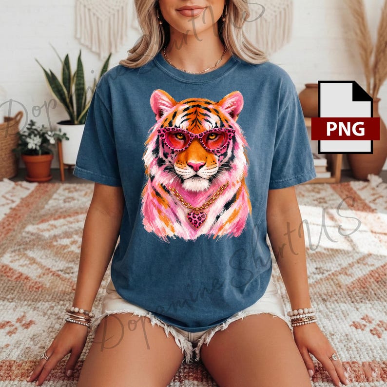 May include: A blue short-sleeve t-shirt featuring a graphic of a tiger wearing pink leopard-print glasses and a gold chain with a heart-shaped pendant. The tiger illustration incorporates vibrant shades of pink, orange, and white.
