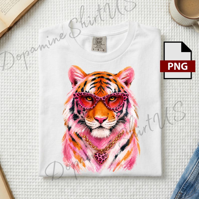 May include: A white t-shirt featuring a graphic illustration of a tiger wearing pink leopard-print glasses and a gold chain with a matching pink leopard-print heart pendant. The tiger has orange and pink fur with black stripes.