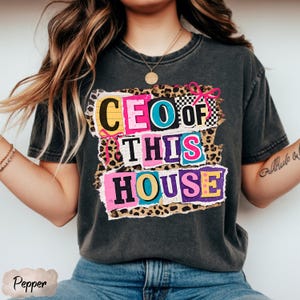 May include: Dark gray t-shirt with the text "CEO OF THIS HOUSE" in colorful, block letter style. The text is layered over a leopard print and checkered background, with pink bow accents. The shirt is a casual wear item.