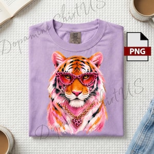 May include: A light purple t-shirt featuring a graphic illustration of a tiger wearing pink leopard-print glasses and a gold chain with a heart-shaped pendant. The shirt has a crew neckline and a Comfort Colors brand label on the inner collar.