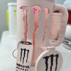 Könnte beinhalten: Ein hellrosa Becher, Schlüsselanhänger und Ornament mit dem Monster Energy-Logo. Der Becher hat einen Henkel und ausgeschnittene Krallen. Der Schlüsselanhänger hat die Form einer Dose und das Ornament ist rund. Das Monster Energy-Logo ist schwarz.