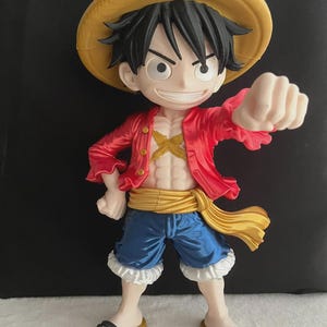 Può includere: Una statuetta stilizzata di un personaggio anime che raffigura una giovane persona con capelli neri, un cappello di paglia giallo, una camicia rossa sbottonata, pantaloncini blu e una fascia dorata. La figura ha una testa grande e un petto definito.