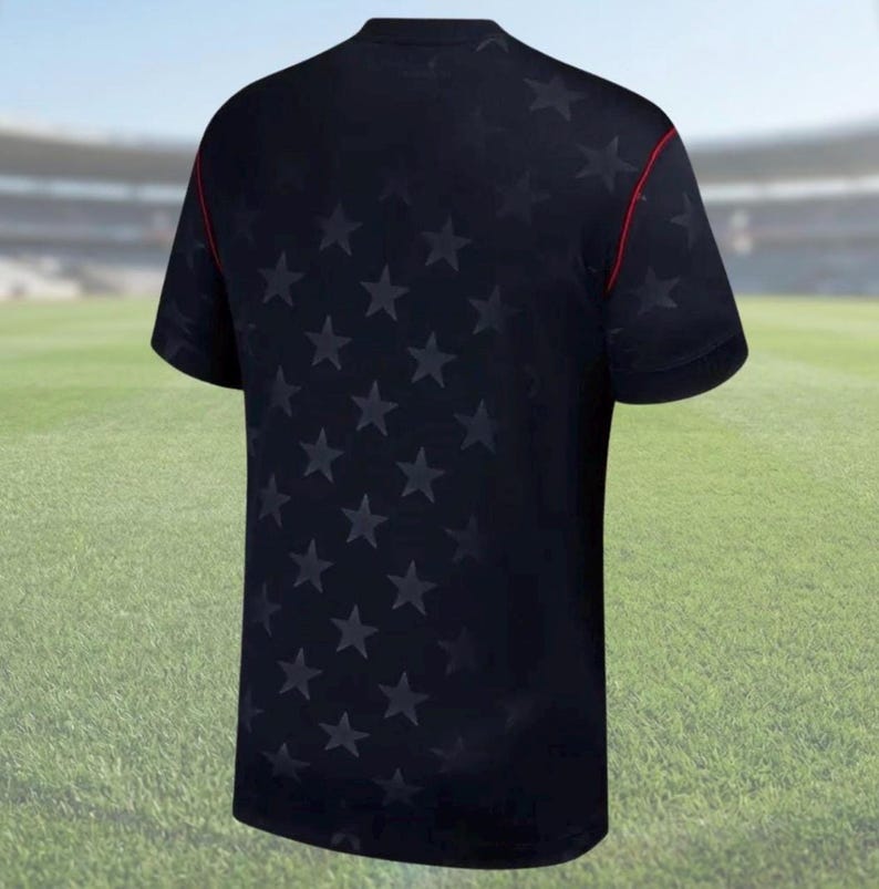 K&ouml;nnte beinhalten: Marineblaues Kurzarm-T-Shirt mit einem Muster aus dunkelblauen Sternen. Das Shirt hat einen Rundhalsausschnitt und rote Paspeln entlang der Schultern. Der Hintergrund zeigt ein gr&uuml;nes Feld und ein Stadion.