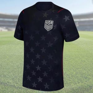 K&ouml;nnte beinhalten: Ein marineblaues T-Shirt mit einem Muster aus dunklen Sternen. Das Shirt hat ein wei&szlig;es und schwarzes USA-Wappen auf der Brust. Rote Akzente sind auf den Schultern und der Seite zu sehen.