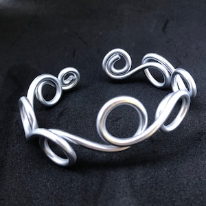 May include: Silver metal bracelet with a swirling, organic design. The bracelet is made of a single piece of wire that is twisted and shaped into a series of loops and curves.
