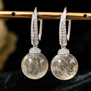 May include: A pair of drop earrings featuring spherical rutilated quartz gemstones with golden needle-like inclusions. The silver-toned lever-back hooks are encrusted with small clear stones, connecting to a pavé-set cap atop each quartz sphere.