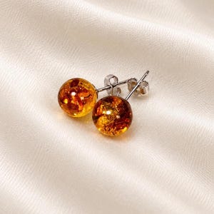 May include: A pair of stud earrings featuring round, polished amber spheres with deep orange and golden internal inclusions. The spherical amber stones are securely mounted on silver-toned metal posts with matching butterfly-style earring backs.