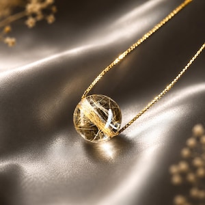 May include: A gold chain necklace featuring a spherical rutilated quartz pendant. The clear, round gemstone contains visible golden needle-like inclusions throughout its interior, suspended from a delicate, polished gold-toned box chain.