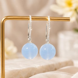 Peut inclure: Une paire de boucles d'oreilles pendantes en argent avec un fil incurvé fin incrusté de petites pierres claires. Chaque boucle d'oreille suspend une perle lisse, ronde et bleu clair qui pend verticalement au bas de la structure en argent.