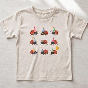 Könnte beinhalten: Ein hellbeiges T-Shirt mit kurzen Ärmeln und einem grafischen Aufdruck von neun Marienkäfern in einem Raster. Jeder Marienkäfer ist mit bunten Partyhüten, herzförmigen Sonnenbrillen oder einem gelben Luftballon als festliches Accessoire verziert.