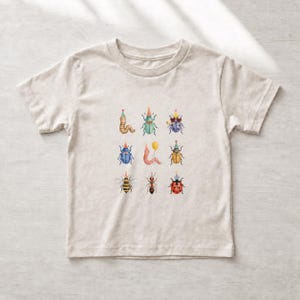 May include: A light beige short-sleeve t-shirt featuring a central graphic print of nine various insects and worms, each wearing a colorful patterned party hat. The illustrations include a ladybug, bee, ant, and other beetles in watercolor style.
