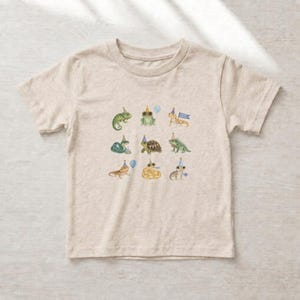 Può includere: Una t-shirt a maniche corte color beige mélange con una stampa grafica centrale raffigurante nove diversi rettili e anfibi, tra cui un camaleonte, una rana, una tartaruga e una lucertola, ognuno con un cappellino da festa e un palloncino.