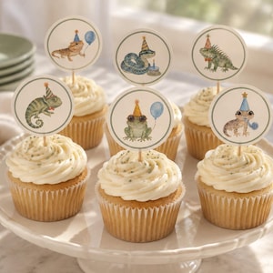 Pode incluir: Seis cupcakes dispostos num suporte branco, cada um com um topo de papel circular apresentando uma ilustração em aguarela de um réptil ou anfíbio com um chapéu de festa, incluindo um camaleão, um sapo, uma cobra e um lagarto.