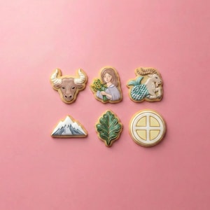 May include: Six decorated sugar cookies arranged in two rows. The top row features a brown bull head, a person holding wheat, and a goat-fish creature. The bottom row includes a mountain peak, a green leaf, and a circular emblem with a cross design.
