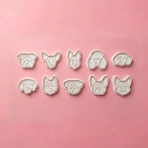 May include: A set of ten white cookie cutters, each shaped like the head of a different dog breed. The cutters are arranged in two horizontal rows of five, featuring detailed embossed facial features for each canine, including ears, eyes, and noses.