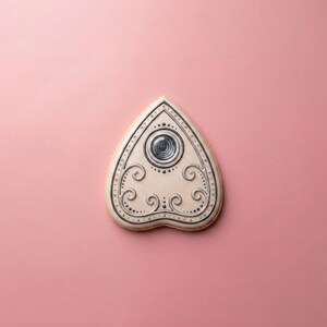 May include: A cream-colored, heart-shaped Ouija board with black and silver decorative accents. The board features a central circular design and ornate scrollwork. The background is a solid pink.
